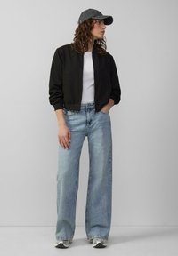 Zwarte cropped bomberjack, witte t-shirt, lichtblauwe wijde jeans, grijze pet en witte sneakers tegen een neutrale achtergrond.