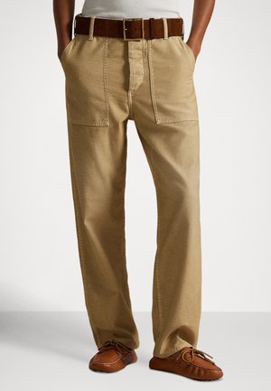Khaki wijde broek met twee voorzakken en een ritssluiting, gecombineerd met een bruine leren riem en tan mocassinachtige schoenen.