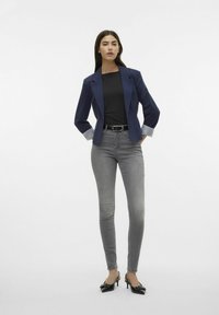 Marinblå blazer med randiga manschetter, svart topp, grå slim fit jeans och svarta klackskor. Släta ytor och skräddarsydd design. Enfärgad bakgrund.