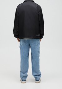 Czarna nylonowa kurtka z kołnierzykiem, w zestawieniu z jasnoniebieskimi dżinsowymi spodniami cargo z bocznymi kieszeniami, noszona z białymi trampkami.