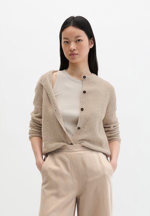 Frau mit langen schwarzen Haaren, die ein beiges, geripptes Tanktop, eine strukturierte beigefarbene Strickjacke mit Knöpfen und hoch taillierte beige Hose trägt, mit den Händen in den Taschen.