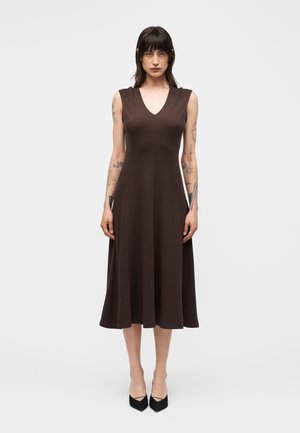 Kobieta o ciemnych włosach i tatuażach, ubrana w brązową sukienkę midi bez rękawów i czarne szpiczaste szpilki, stojąca na jednolitym jasnym tle.