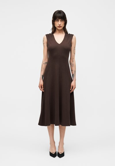 Femme aux cheveux foncés et tatouages, portant une robe midi marron sans manches et des talons pointus noirs, debout devant un fond clair uni.