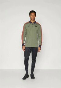 Camisa polo de manga longa verde com listas vermelhas nos ombros e punhos pretos, combinada com calças pretas com listas brancas laterais.