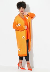 Dlouhý oranžový cardigan s bílými květinovými aplikacemi, kombinovaný s oranžovým topem a černými legínami. Má uvolněný střih a vroubkované manžety.