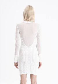 Robe blanche à manches longues ; partie supérieure transparente et moitié inférieure en maille côtelée texturée. Caractérisée par une découpe profonde dans le dos et une silhouette ajustée.