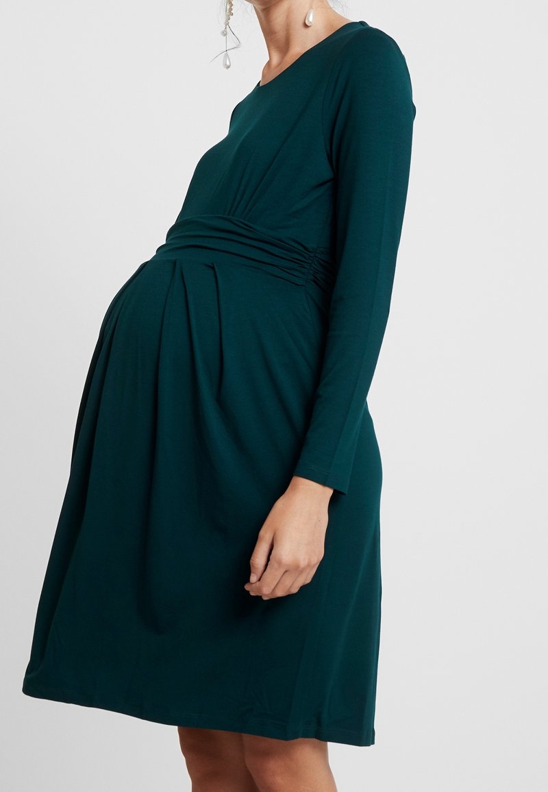 Mörk teal gravidklänning, lång ärm, rynkad midja, knälång, tillverkad av mjukt tyg med veckad design.