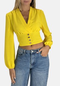 Gul langærmet crop top med V-udskæring og knaplukning, kombineret med højtaljede blå denimjeans. Glat stof og tætsiddende design.