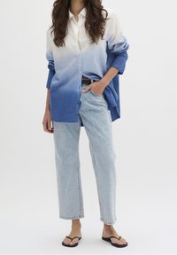 Kvinna iklädd en blårandig skjorta med ombré-effekt, ljusblå högmidjade raka jeans, svart bälte och svarta flip-flops, stående mot en vit bakgrund.