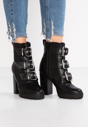 Bottines en cuir noir avec trois brides à boucle argentée, talons épais, fermetures éclair latérales, portées avec un jean bleu à ourlet effiloché.