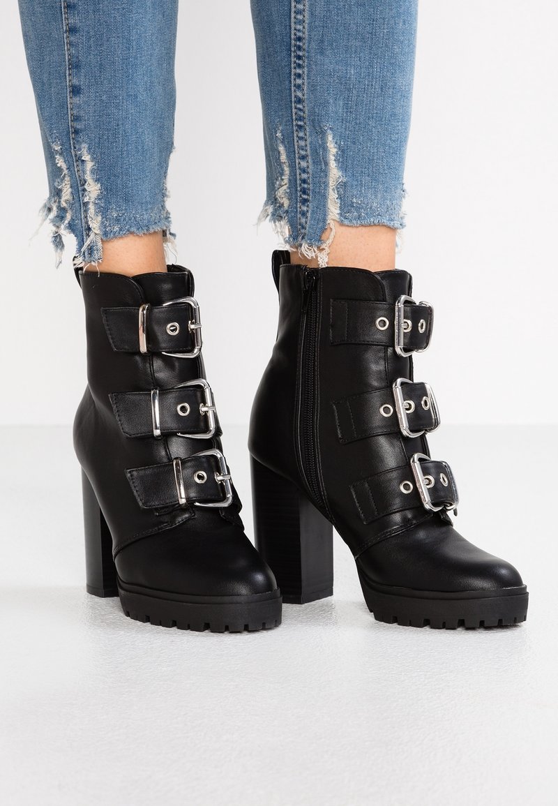 Bottines en cuir noir avec trois brides à boucle argentée, talons épais, fermetures éclair latérales, portées avec un jean bleu à ourlet effiloché.