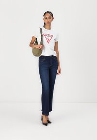 T-shirt blanc avec logo en forme de triangle rouge, jean droit bleu foncé et talons noirs. Le mannequin tient un sac à bandoulière vert olive.