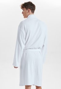 Albornoz blanco con textura de waffle, de manga larga, con cinturón y corte hasta la rodilla. Presenta un cuello de chal y detalles acanalados en los puños.