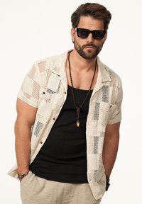 Weißes kurzärmeliges Offenstrickshirt mit einem schwarzen Tanktop darunter, beigen strukturierten Hosen, Sonnenbrille und einer geschichteten Halskette.