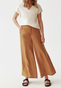 Femme portant un pantalon large kaki, un haut crème à manches courtes en V, et des sandales noires à bout ouvert avec des boucles dorées, debout avec une main dans la poche.