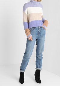 Pull en tricot avec un col haut, présentant des rayures horizontales en lavande, blanc et beige. Associé à un jean bleu clair et des bottes à talons noires.