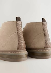 Tan-farbene Wildleder-Ankle-Boots mit strukturierten braunen Einsätzen, einer Gummisohle und gestreiften Details an der Unterseite. Mit einer Schlaufe an der Rückseite für einfaches An- und Ausziehen.