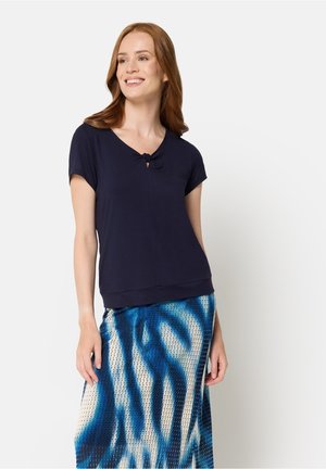 Lächelnde Frau mit roten Haaren, die ein schwarzes kurzärmeliges Oberteil und einen blau gemusterten Rock trägt, steht vor einem weißen Hintergrund.