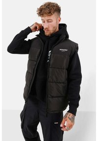 Zwarte puffer vest met een ritssluiting, voorzien van een lichtgewicht, gewatteerd ontwerp, gedragen over een zwarte hoodie. Grijze branding op de borst.
