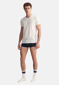 Weißes Baumwoll-V-Ausschnitt-T-Shirt, kombiniert mit marineblauen Unterhosen und weißen Socken mit marinen Streifen. Das Model steht aufrecht und schaut geradeaus.