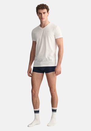 Weißes Baumwoll-V-Ausschnitt-T-Shirt, kombiniert mit marineblauen Unterhosen und weißen Socken mit marinen Streifen. Das Model steht aufrecht und schaut geradeaus.