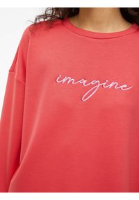 Koralowa bluza z haftowanym różowym napisem "imagine" kursywą. Charakteryzuje się luźnym krojem i miękką fakturą oraz klasycznym okrągłym dekoltem.