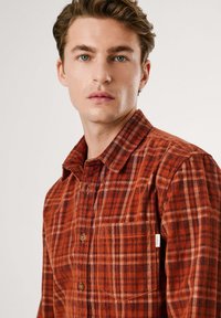 Camicia di flanella a quadri rossi e arancioni, con colletto a camicia, tasca frontale e bottoni marroni. Tessuto morbido con una vestibilità rilassata.