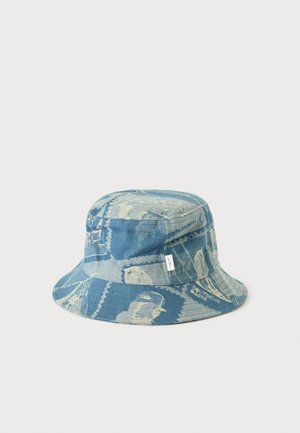 Chapeau bob en denim bleu avec des motifs floraux et géométriques légers et une petite étiquette blanche sur le côté, sur un fond clair uni.