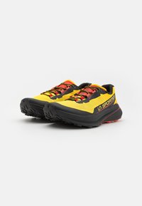 Scarpe sportive gialle con accenti neri e rossi, superiore in mesh testurizzato e suola nera robusta per una buona aderenza. Sistema di allacciatura per una vestibilità sicura.