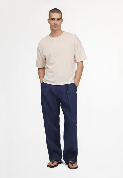 Homme aux cheveux courts portant un t-shirt beige oversize, un pantalon large bleu marine et des tongs noires, debout les mains dans les poches.