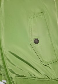 Giacca verde con una superficie liscia e lucida, chiusura con zip e una tasca visibile con dettaglio a pulsante circolare.