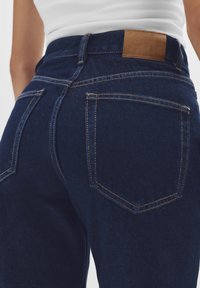 Jeans in denim blu scuro con vita alta, dotati di due tasche posteriori e una patch in pelle marrone con logo impresso sulla vita.