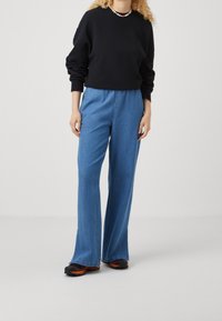 La persona indossa un maglione nero a maniche lunghe, jeans a vita alta e gamba larga blu, sneakers nere e arancioni, e una collana di perline, in piedi su un pavimento bianco.
