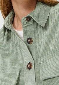 Green corduroy jacket featuring a pointed collar, large buttons, and textured vertical ridges. Includes front pockets.