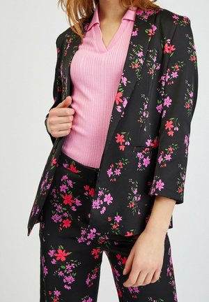Femme portant un blazer et un pantalon noirs avec des motifs floraux roses et rouges, associés à un haut côtelé rose clair à col en V.
