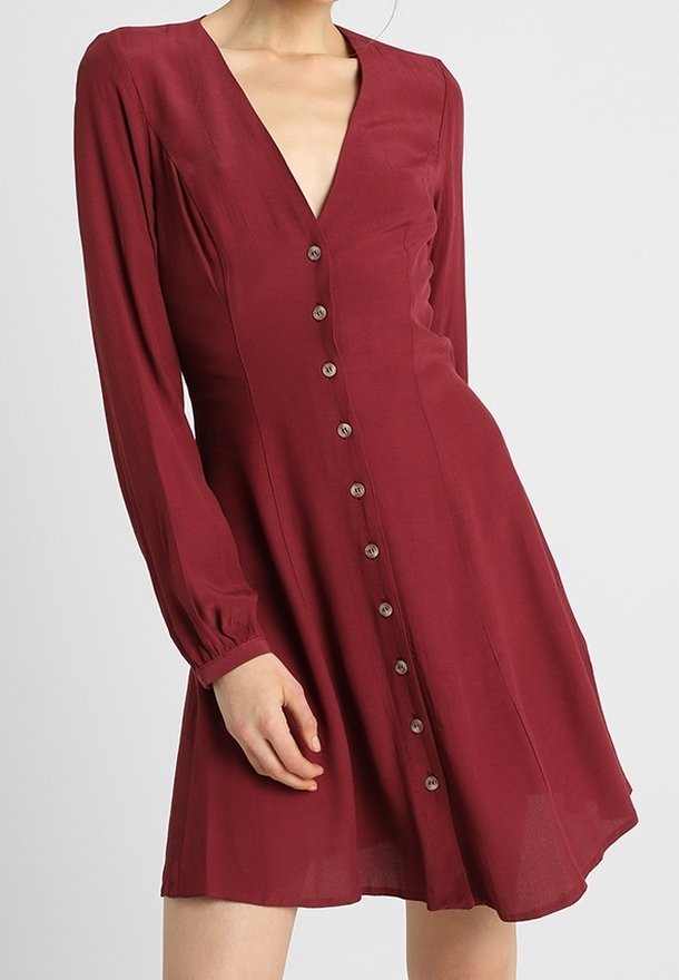 Femme portant une robe rouge foncé à manches longues, boutonnée, avec un décolleté en V et une jupe légèrement évasée, sur un fond uni.