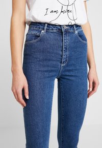 Modré džíny s vysokým pasem vyrobené z denimu, s úzkým střihem, předními kapsami a zapínáním na knoflík s kontrastním prošíváním.