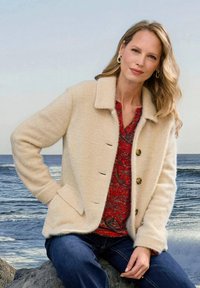 Beige wollen jas met een kraag, twee voorkanten zakken en vijf knopen. Draagt over een rood patroon top en gecombineerd met blauwe jeans. Getextureerd materiaal.