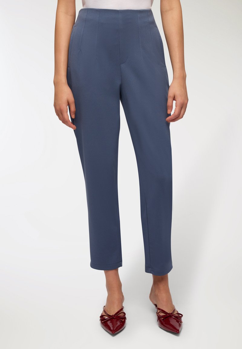 Dopasowane niebieskie spodnie mają wysoki stan, proste nogawki i boczne kieszenie. Założone z czerwonymi błyszczącymi balerinami zakończonymi na szpic, zdobionymi kokardkami.