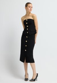 Černé bezramenné šaty se strukturovaným korzetem, peplum pasem, předním zapínáním na zlaté knoflíky a přiléhavou sukní. Spárované s černými lodičkami s špičatou špičkou.