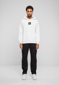 Hvid hættejakke med en sort logo-patch, kombineret med sorte cargo-bukser med sidelommer og hvide sneakers. Glat tekstur med en afslappet pasform.