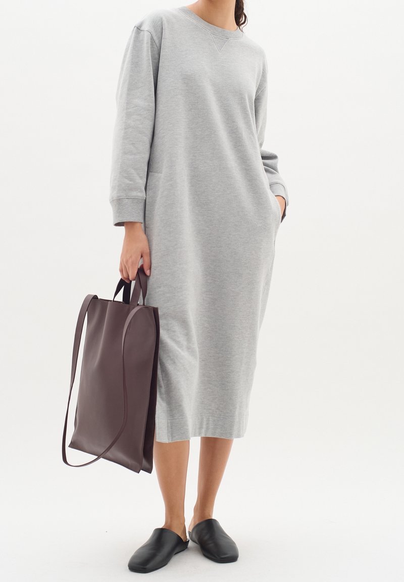Graues Sweatshirtkleid mit langen Ärmeln, Seitentaschen und Rundhalsausschnitt, kombiniert mit einer dunkelbraunen Tasche und schwarzen Slippern.