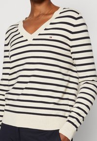 Pull à col en V rayé crème et marine, avec manches longues, poignets côtelés et un petit logo sur la poitrine. Texture douce.