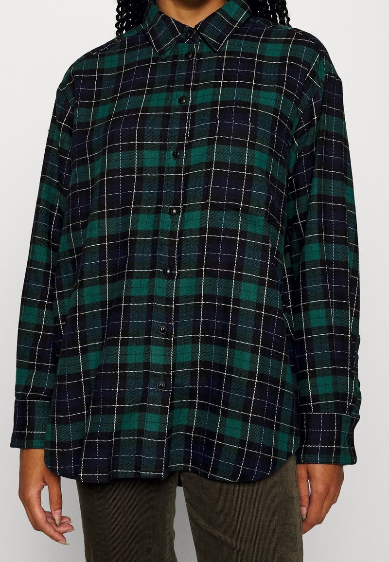 Chemise en flanelle à carreaux vert foncé et bleu marine, avec des coutures blanches, un col boutonné et des manches longues. Tissu en coton texturé.