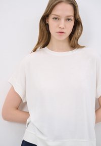 Bílý krátký rukáv s volným střihem a kulatým výstřihem. Hladká látka s mírným leskem. Modelka má ruce na bocích na neutrálním pozadí.