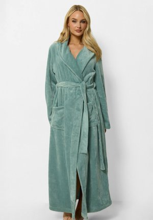 Femme portant un long peignoir doux bleu-vert avec une ceinture et deux poches avant, debout devant un fond blanc uni.
