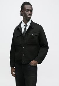 Čierna rifľová bunda s golierom, dvoma náprsnými vreckami a jemným logom. Nosená na bielej pásikovanej košeli a čiernej kravate.