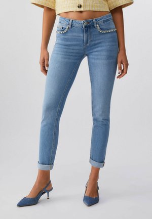 Donna che indossa jeans skinny azzurri chiari con risvolti arrotolati e dettagli in strass sulle tasche, abbinati a tacchi slingback blu e top giallo corto.