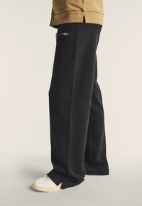 Pantalones negros de tela suave, con un corte relajado, abertura lateral en el dobladillo y un pequeño logo "FREDDY" en la parte delantera. Combínalos con zapatillas blancas.