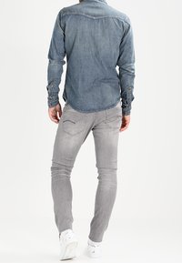 Mans denimskjorta, blå, långärmig, med knapp-down krage, tillsammans med grå slim-fit jeans, med bakfickor och vita sneakers.